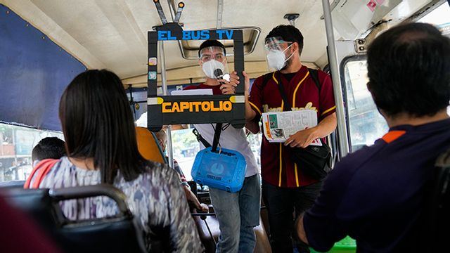 Juan Pablo Lares, right, holds a cardboard frame in front of his associate Maximiliano Bruzual who reads their newscast ”El Bus TV Capitolio” to commuters on a bus in Caracas, Venezuela, Saturday, July 31, 2021. Two decades of governments that see the press as an enemy have pushed Venezuelan journalists to find alternative ways to keep citizens informed. Photo: Ariana Cubillos/AP