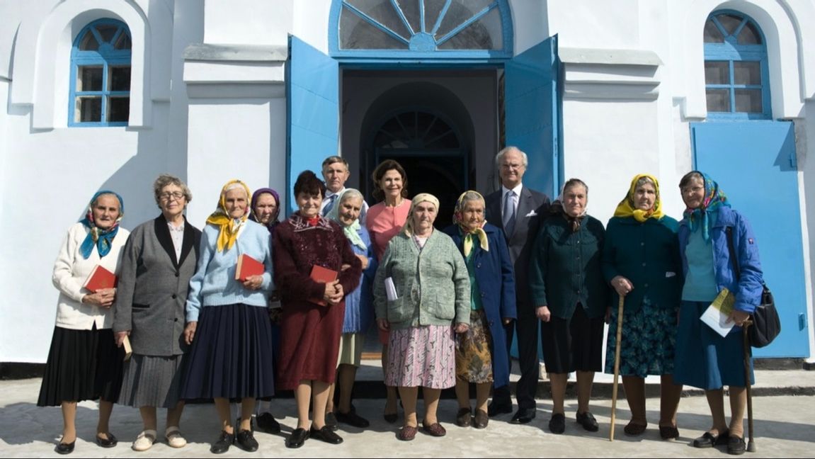 Sveriges kung och drottning framför kyrkan i Gammalsvenskby vid statsbesök 2008, omgivna av några av de sista svensktalande i byn. Foto: Henrik Montgomery/TT